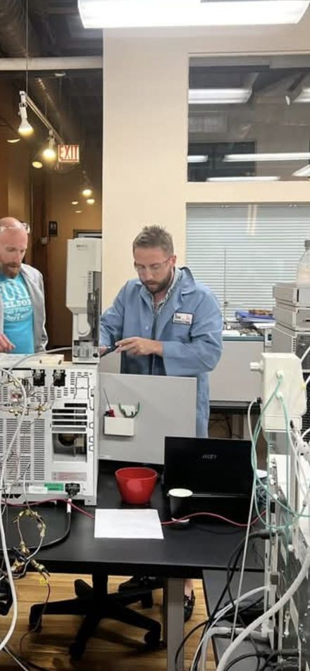 Rory Safir in a lab coat and safety glasses operating gas chromatography instrumentation at the Axion bench